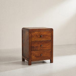 Könnte beinhalten: Ein Nachttisch aus Holz mit drei Schubladen in warmbrauner Farbe. Der Nachttisch hat eine abgerundete Oberseite und kleine, dunkle Knöpfe an jeder Schublade. Die Holzmaserung ist sichtbar und verleiht dem Möbelstück Textur. Ein klassisches Schlafzimmermöbel.
