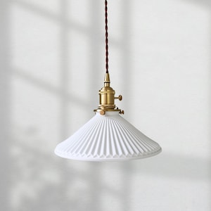Könnte beinhalten: Weiße Pendelleuchte mit plissiertem Schirm und Messingakzenten. Die Leuchte hängt an einem braunen Kabel. Das moderne Design eignet sich für Küche oder Esszimmer. Das Logo "ev lighting" befindet sich unten.