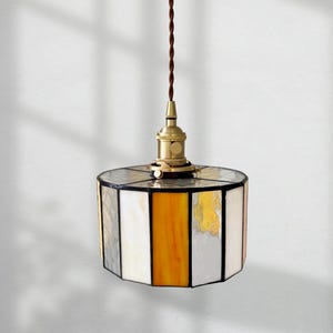 Könnte beinhalten: Hängeleuchte mit einem Lampenschirm aus Buntglas. Der Schirm hat eine geometrische Form mit weißen, grauen, gelben und goldenen Paneelen. Die Leuchte hat eine messingfarbene Fassung und ein braunes Kabel. Das Logo "ev lighting" befindet sich unten.