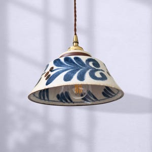 Könnte beinhalten: Eine Keramik-Pendelleuchte mit weißem Sockel und blauem Blumendesign. Die Leuchte hat ein braunes Kabel und eine messingfarbene Oberseite. Die Glühbirne ist im Inneren des Schirms sichtbar. Das Logo "ev lighting" befindet sich unten.