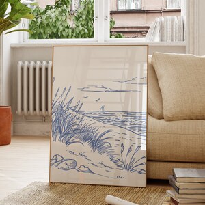 Peut inclure: Une affiche encadrée représentant une scène côtière bleue et blanche. L'œuvre est exposée sur un tapis beige dans un salon lumineux avec une chaise en bois, une plante en pot et un canapé beige. La pièce a une fenêtre.