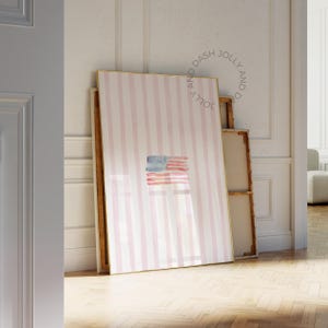 Könnte beinhalten: Ein gerahmter Kunstdruck mit einer Aquarelldarstellung der amerikanischen Flagge. Der Druck hat einen rosa-weiß gestreiften Hintergrund und einen goldenen Rahmen. Die Flagge ist in Blau- und Rottönen gemalt.
