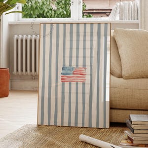 Könnte beinhalten: Ein gerahmter Aquarelldruck mit der amerikanischen Flagge. Die Flagge ist in Rot, Weiß und Blau gemalt, vor einem Hintergrund aus hellblauen und cremeweißen Streifen. Das Kunstwerk wird in einem Raum mit natürlichem Licht ausgestellt.