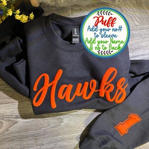 May include: A black crewneck sweatshirt featuring the word Hawks in raised orange puff lettering across the chest and the number 1 in orange on the sleeve. A circular graphic overlay includes the text Puff, Add your no# to sleeve, Add your Name & no to Back.