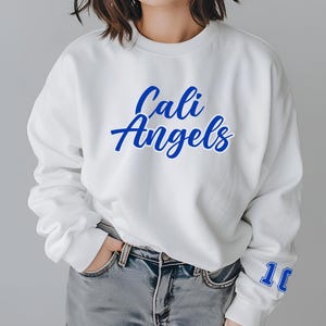 Puede incluir: Sudadera blanca de cuello redondo con las palabras "Cali Angels" en escritura azul en el pecho. El n&uacute;mero "10" est&aacute; impreso en azul en la manga derecha. La persona lleva vaqueros azules.