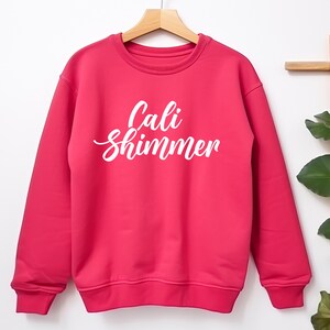 Puede incluir: Una sudadera rosa brillante con la inscripci&oacute;n blanca "Cali Shimmer". La prenda est&aacute; colgada de una percha de madera. La parte superior de manga larga est&aacute; colocada sobre un fondo blanco, con un vistazo de follaje a la derecha.