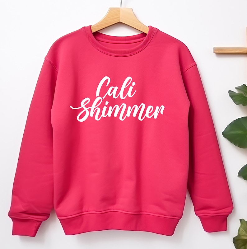 Puede incluir: Una sudadera rosa brillante con la inscripci&oacute;n blanca "Cali Shimmer". La prenda est&aacute; colgada de una percha de madera. La parte superior de manga larga est&aacute; colocada sobre un fondo blanco, con un vistazo de follaje a la derecha.