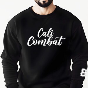 Puede incluir: Sudadera negra con el texto blanco "Cali Combat" en cursiva en el pecho. El n&uacute;mero "8" est&aacute; en la manga derecha. La sudadera es de cuello redondo.