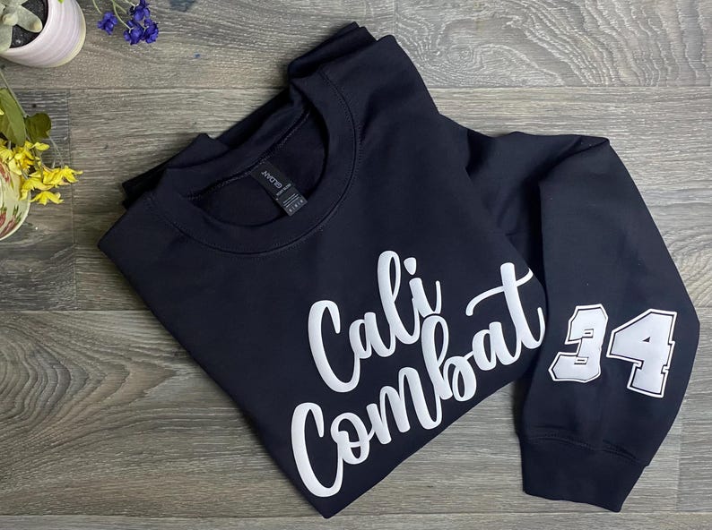 Puede incluir: Una sudadera azul marino con las palabras "Cali Combat" en escritura blanca en la parte delantera. El n&uacute;mero "34" est&aacute; en la manga derecha en blanco con un contorno negro. La sudadera est&aacute; doblada sobre una superficie de madera.