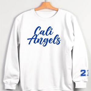Puede incluir: Sudadera blanca con las palabras "Cali Angels" en escritura azul en el pecho. El n&uacute;mero "21" est&aacute; impreso en azul en la manga izquierda. La sudadera est&aacute; colgada en una percha de madera.