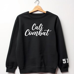 Puede incluir: Sudadera negra con el texto blanco "Cali Combat" en el pecho. El n&uacute;mero "51" est&aacute; en la manga izquierda en blanco. La sudadera est&aacute; colgada en una percha de madera.