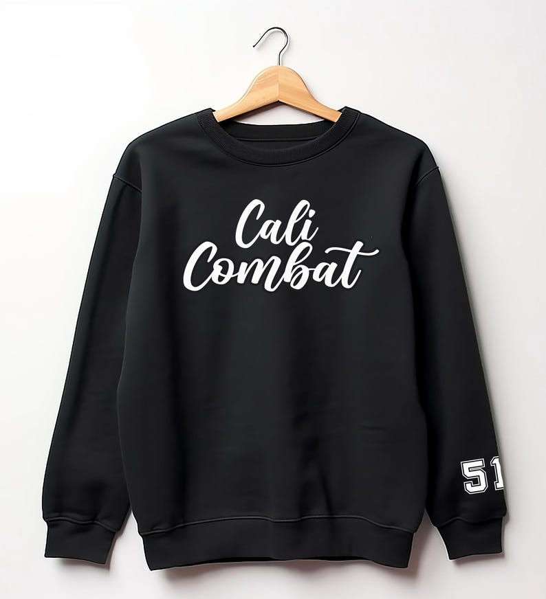 Puede incluir: Sudadera negra con el texto blanco "Cali Combat" en el pecho. El n&uacute;mero "51" est&aacute; en la manga izquierda en blanco. La sudadera est&aacute; colgada en una percha de madera.