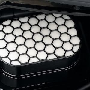 Puede incluir: Filtro de aire negro con un patrón de panal. El filtro tiene un marco negro y un interior blanco y poroso. La imagen es un primer plano, que destaca el diseño geométrico y la textura del filtro.