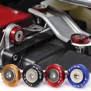 Può includere: Primo piano di diversi bulloni automobilistici in oro, nero, rosso e blu. Ogni bullone ha una testa circolare con il testo "PWM JDM" e caratteri giapponesi. I bulloni sono installati su un componente del veicolo.