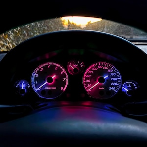 May include: A car dashboard with illuminated gauges. The speedometer and tachometer glow with pink and blue lights. The speedometer reads up to 240 km/h. The tachometer shows up to 8 x1000/min. The steering wheel is visible in the foreground.