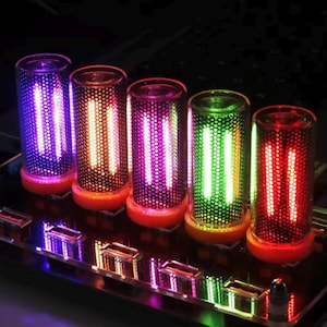 Può includere: Cinque display a tubo nixie cilindrici disposti in fila su una scheda a circuito stampato. Ogni tubo presenta un rivestimento in rete metallica nera perforata, una parte superiore in vetro trasparente e una base arancione. I tubi emettono una luce brillante viola, arancione, rosa, verde e rossa.