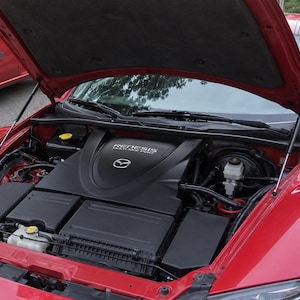 May include: The open bonnet of a red car reveals a black engine cover with the text RENESIS MULTI-SIDE PORT and a Mazda logo. The engine bay contains various black plastic components, fluid reservoirs with yellow caps, and metal mechanical parts.