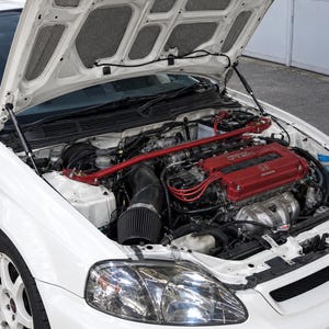 Puede incluir: Un Honda Civic blanco con el capó abierto, que muestra una tapa de válvulas de motor Honda VTEC roja, una barra de refuerzo roja y un filtro de aire cónico negro. El compartimento del motor presenta varios tubos negros y colectores.