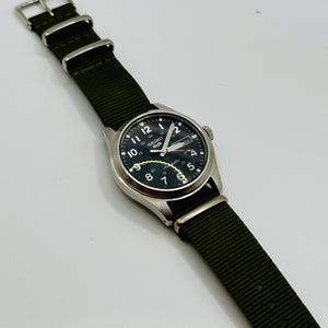 Peut inclure: Une montre-bracelet argentée avec un cadran noir et un bracelet en nylon vert. Le cadran affiche des chiffres et des aiguilles blancs, avec la marque "SEIKO". Le bracelet est vert olive foncé.