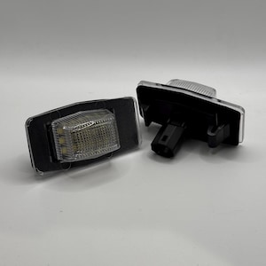Puede incluir: Dos luces LED para matrícula. Cada luz tiene una lente rectangular transparente con una rejilla de pequeñas bombillas LED. Las luces están en una carcasa negra con un conector negro. Una luz está separada, mostrando el conector.