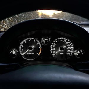 May include: A black car dashboard with white gauges. The speedometer reads 0 km/h and the tachometer reads 0 rpm. The gauges are backlit with a white light.