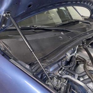 Peut inclure: Un capot de voiture bleu est ouvert, révélant le compartiment moteur. Des vérins noirs maintiennent le capot en place. Le moteur est visible, avec divers tuyaux et composants. La carrosserie de la voiture est bleu métallisé.