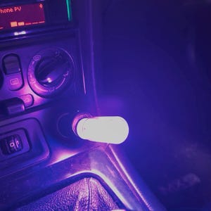 Puede incluir: Detalle interior del coche con una luz blanca brillante que emana de un cargador de coche. El salpicadero presenta una pantalla de radio y controles de aire acondicionado, todo en un tono púrpura. La imagen captura el interior del coche.