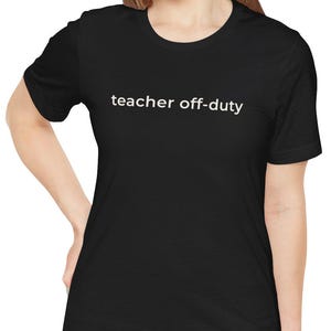 May include: Black t-shirt with the text "teacher off-duty" in white, centered on the chest. The shirt is a short-sleeved crew neck style. The model is wearing blue jeans.