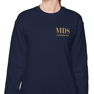 Könnte beinhalten: Marineblaues Sweatshirt mit goldenem Schriftzug "MDS COORDINATOR". Das Sweatshirt hat einen Rundhalsausschnitt. Die Person trägt eine blaue Jeans.