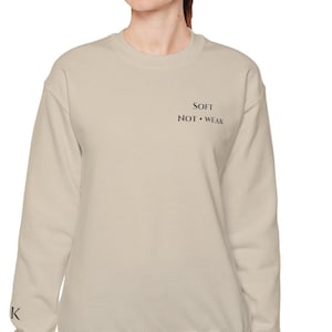 Może przedstawiać: Beżowa bluza z okrągłym dekoltem z czarnym napisem "SOFT NOT WEAK" z przodu. Bluza ma długie rękawy i luźny krój. Litera "K" jest nadrukowana na lewym rękawie.