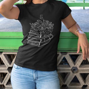 May include: Black t-shirt featuring a white graphic of roses and a butterfly atop a stack of books. The design is a line drawing. The model is wearing blue jeans. The shirt is a crew neck.