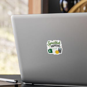 May include: A square sticker featuring the green text Spreadsheet Happens, a white spreadsheet grid with a green X icon, a grey calculator, and a yellow smiley face, all displayed on the lid of a silver laptop computer.