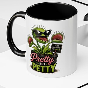 May include: A white ceramic mug with a black interior and handle. The exterior features a graphic of a green Venus flytrap wearing black sunglasses, planted in a black pot. The text reads Pretty BUT PETTY and ZERO TOLERANCE FOR IDIOTS.