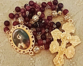 May include: A rosary with deep red and dark purple beads. The rosary features a gold-colored crucifix and a gold-framed oval pendant with a portrait. The beads are strung on a gold-colored chain.