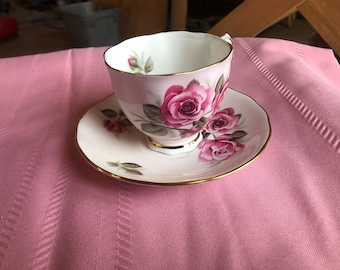 May include: A white teacup and saucer with a pink and gold floral design. The cup and saucer feature a delicate floral pattern with pink roses and green leaves.