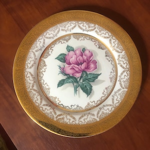 May include: A white porcelain dinner plate with a gold rim and intricate gold design. The center of the plate features a pink rose with green leaves.