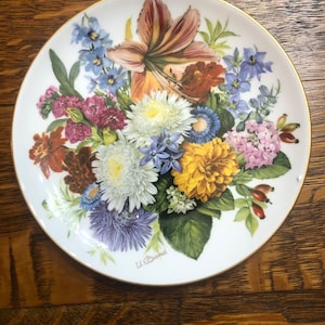May include: A white porcelain plate with a gold rim features a colorful floral bouquet. The bouquet includes lilies, chrysanthemums, and other flowers in shades of pink, purple, yellow, and orange. The plate is signed "U. Band".