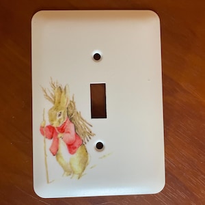 May include: White light switch cover featuring a watercolor illustration of a rabbit wearing a red jacket and carrying a bundle of sticks. The cover has two screw holes and a rectangular opening for the switch.