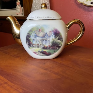 May include: White ceramic teapot with gold accents, featuring a scenic illustration of a house and garden. The lid has the text "Home Is Where The Heart Is". The teapot has a gold handle, spout, and lid finial.