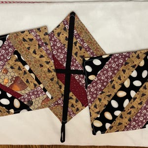 May include: Set of three quilted pot holders with a farm theme. The pot holders feature a patchwork design with egg, chicken, and floral patterns in black, white, brown, and burgundy colors. Each pot holder has a loop for hanging.
