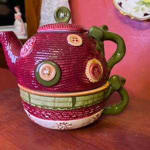 May include: A decorative teapot and teacup set. The teapot is a deep red with a textured knit design and adorned with decorative buttons. The teacup is a matching design with green handles and trim. The set is designed for serving tea.