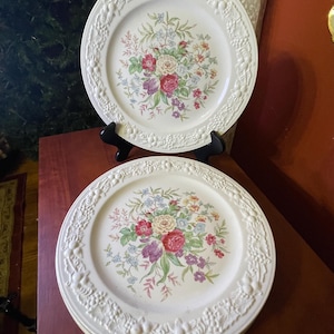 May include: Two white ceramic dinner plates with a floral design. The plates have a raised, textured border with a floral and leaf pattern. The center of the plates features a bouquet of red, pink, purple, and blue flowers.