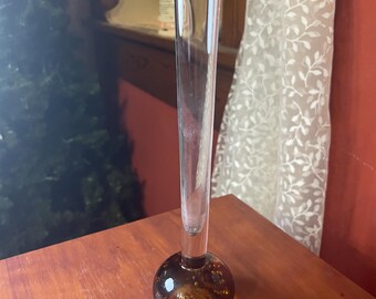 May include: A clear glass bud vase with a round, amber-colored base. The vase has a long, slender neck and a flared top. The base has a textured appearance. The vase is sitting on a wooden surface.