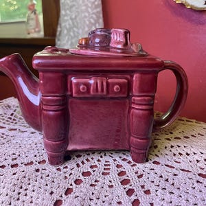 May include: A burgundy ceramic teapot shaped like a dresser, with a spout, handle, and a lid with miniature objects on top. The teapot has a drawer and is set on a cream-colored lace doily.