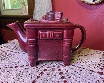 Op de afbeelding: Bordeauxrode keramische theepot in de vorm van een dressoir, met een tuit, handvat en een deksel met miniatuurvoorwerpen erop. De theepot heeft een lade en staat op een crèmekleurig kanten kleedje.