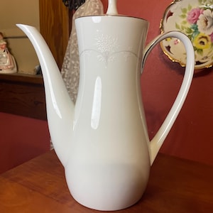 May include: A white porcelain coffee pot with a slender spout and curved handle. The pot features a delicate floral pattern and gold trim. The lid has a small, white finial. The pot is on a wooden surface.