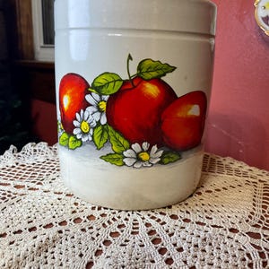 May include: A cylindrical, off-white ceramic canister with a hand-painted design of red apples, green leaves, and white daisies. The canister is likely for kitchen storage and has a glossy finish.