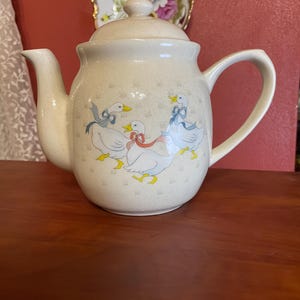 May include: Cream-colored ceramic teapot with a lid and handle. The teapot is decorated with illustrations of geese wearing bows. The background has a subtle floral pattern. A decorative plate with floral designs is visible in the background.