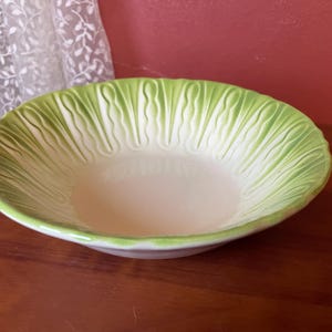 May include: A ceramic serving bowl with a lettuce leaf design. The bowl has a light green rim and a white interior. The bowl is sitting on a wooden surface. The bowl is a decorative piece.