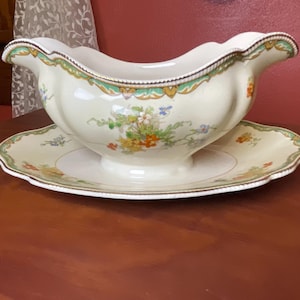 May include: Cream-colored gravy boat and matching saucer with floral and gold trim. The gravy boat has a scalloped rim and a handle. The saucer is round with a scalloped edge. The set is decorated with colorful flowers.